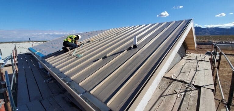 Worker installing metal roofing panels on water facility building with safety harness and scaffolding in place.
