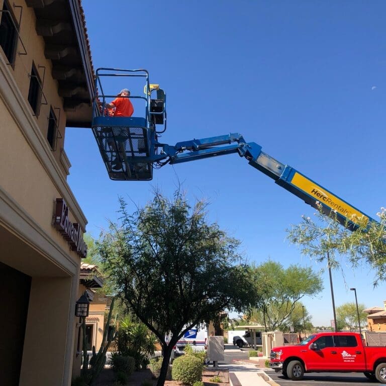 Worker on a boom lift performing fascia remodeling work on the exterior roof overhang of a commercial plaza building under a clear blue sky.