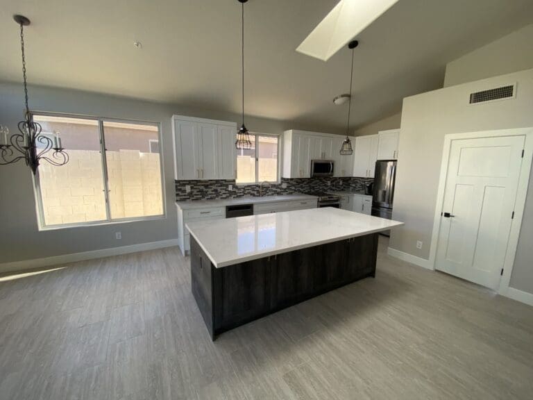 Wide-angle after photo of the North Scottsdale kitchen remodel featuring a bright open-concept layout, oversized quartz island, white shaker cabinets, stainless steel appliances, gray tile flooring, and modern pendant lighting