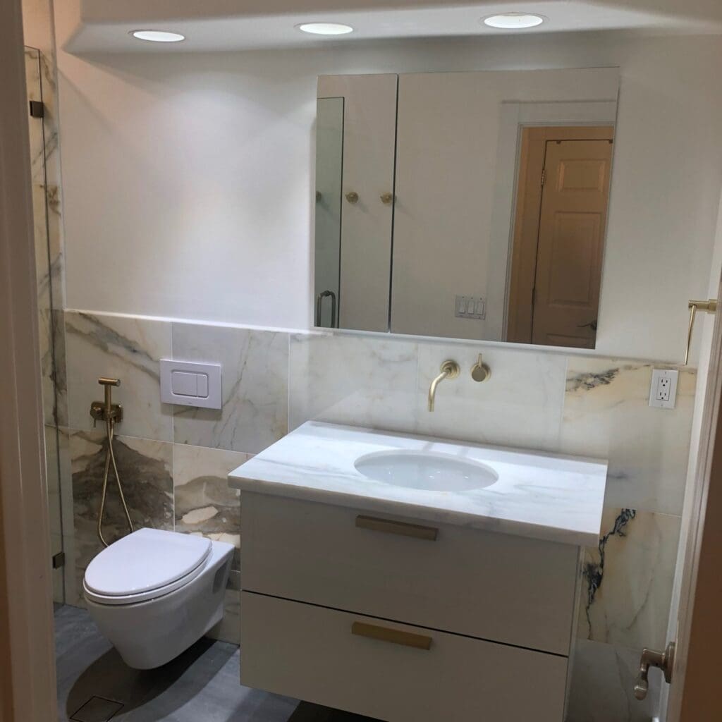 Contemporary bathroom vanity with marble countertop, wall-mounted brushed-gold faucet, large mirrored cabinet, and wall-hung toilet with marble accent tiles.