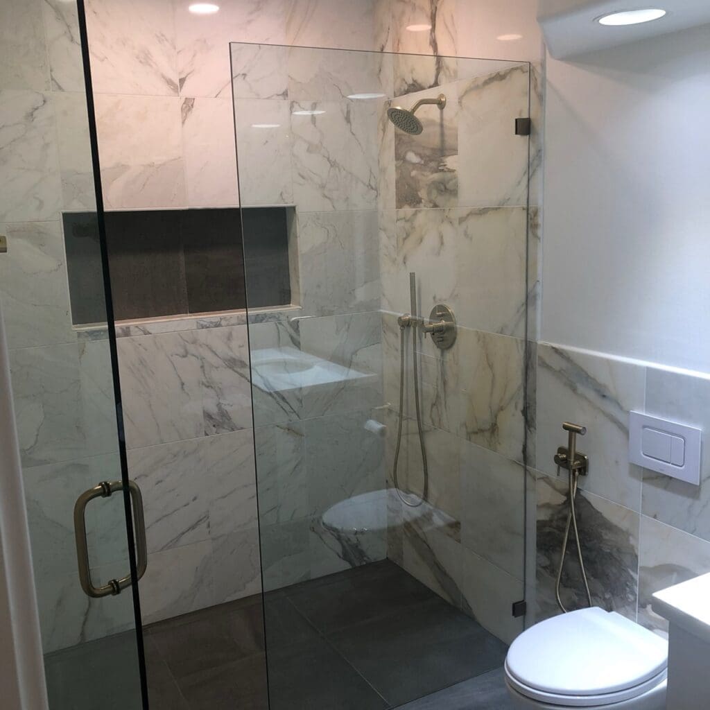 Modern bathroom shower with glass enclosure, large marble-style wall tiles, built-in niche, and brushed-gold shower fixtures.