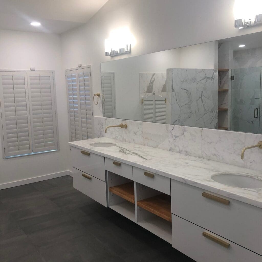 Modern bathroom with a long double-sink vanity, white marble countertop, gold fixtures, large wall mirror, and shuttered windows