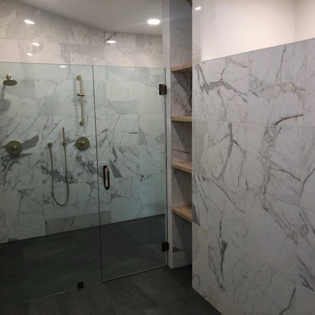 Marble-tiled bathroom with frameless glass shower enclosure and built-in wooden shelving along the adjacent wall