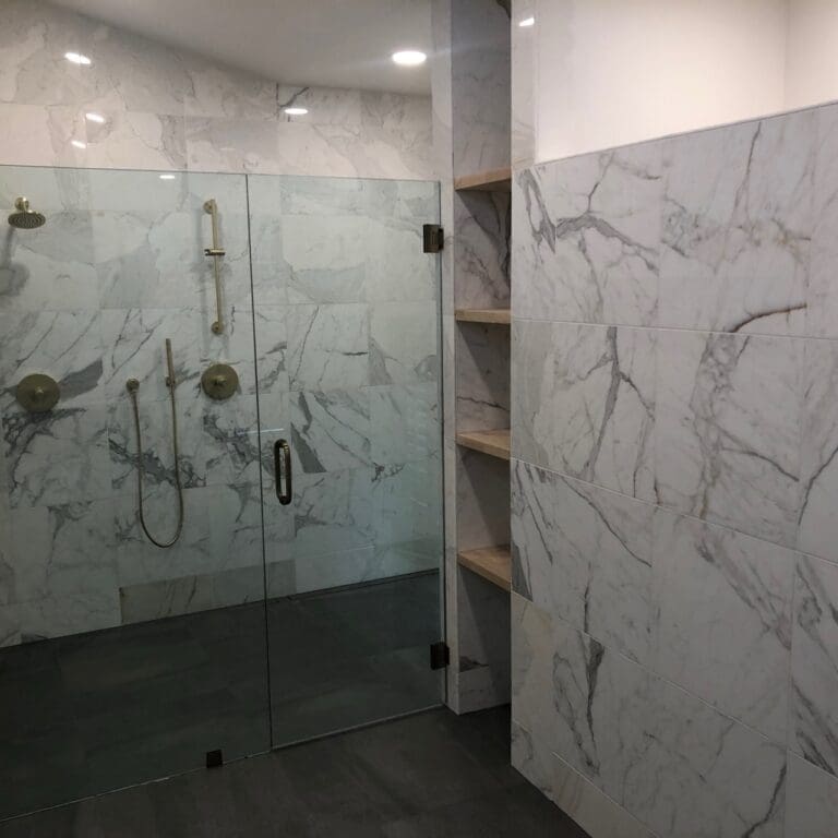 Marble-tiled bathroom with frameless glass shower enclosure and built-in wooden shelving along the adjacent wall