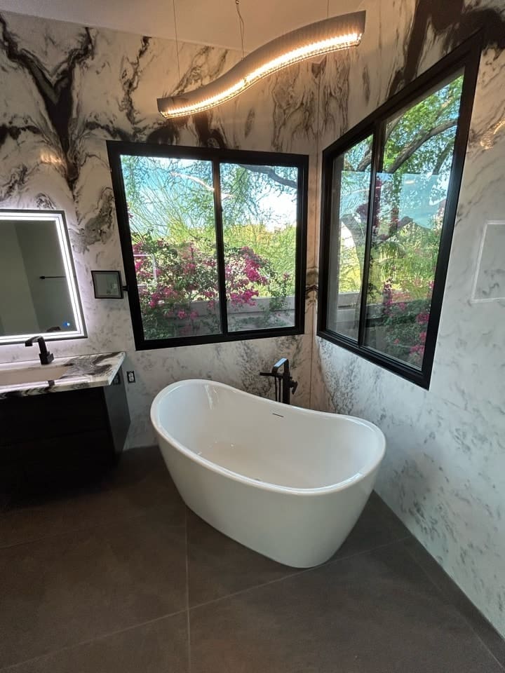 Freestanding white bathtub positioned beneath large corner windows with garden views, set against dramatic marble-patterned walls and a sculptural curved light fixture overhead