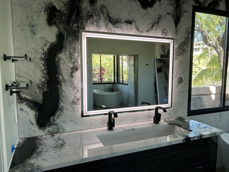 Marble-accent bathroom vanity with a wide backlit mirror, black fixtures, and large windows reflecting a freestanding tub and outdoor greenery
