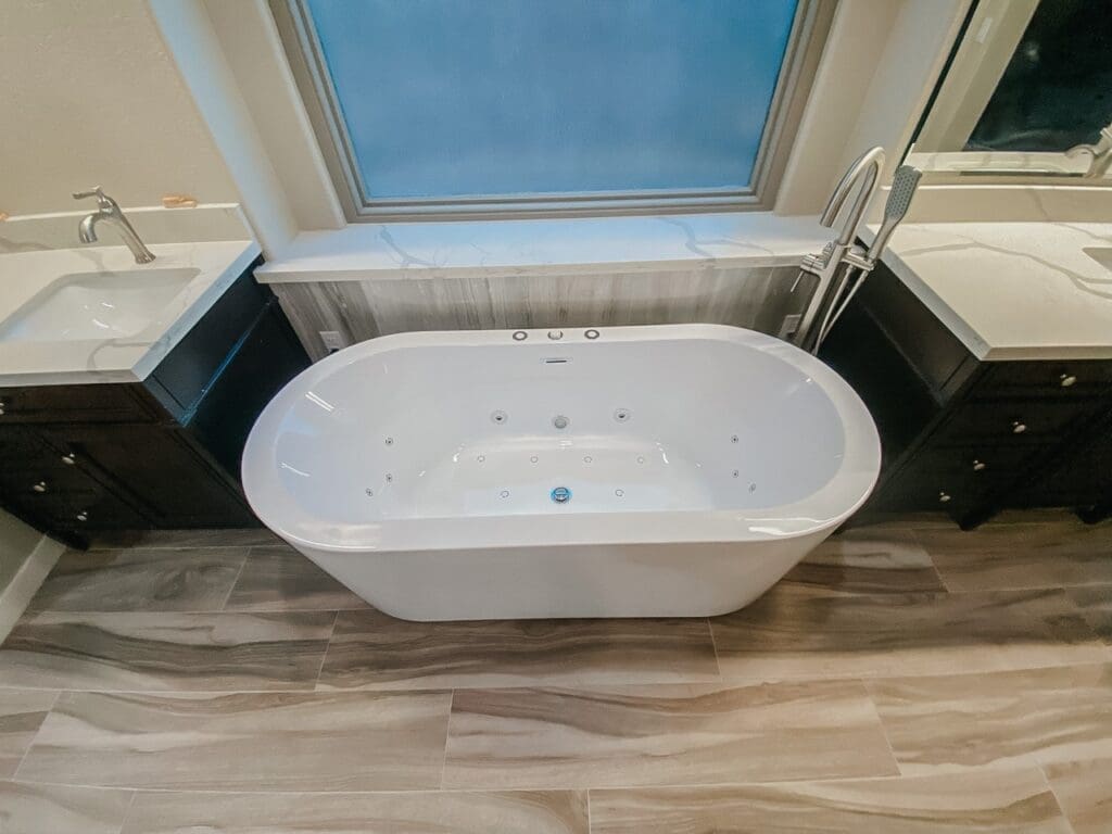 Freestanding soaking tub positioned between two vanities, placed beneath a large frosted window and set on wood-look tile flooring