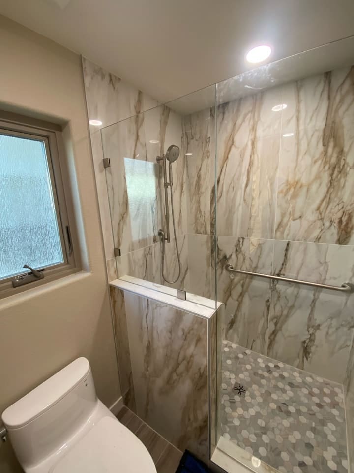 Bathroom with a glass-enclosed shower featuring marble-look tile, a built-in ledge, hexagon floor tile, and a toilet beside a frosted window