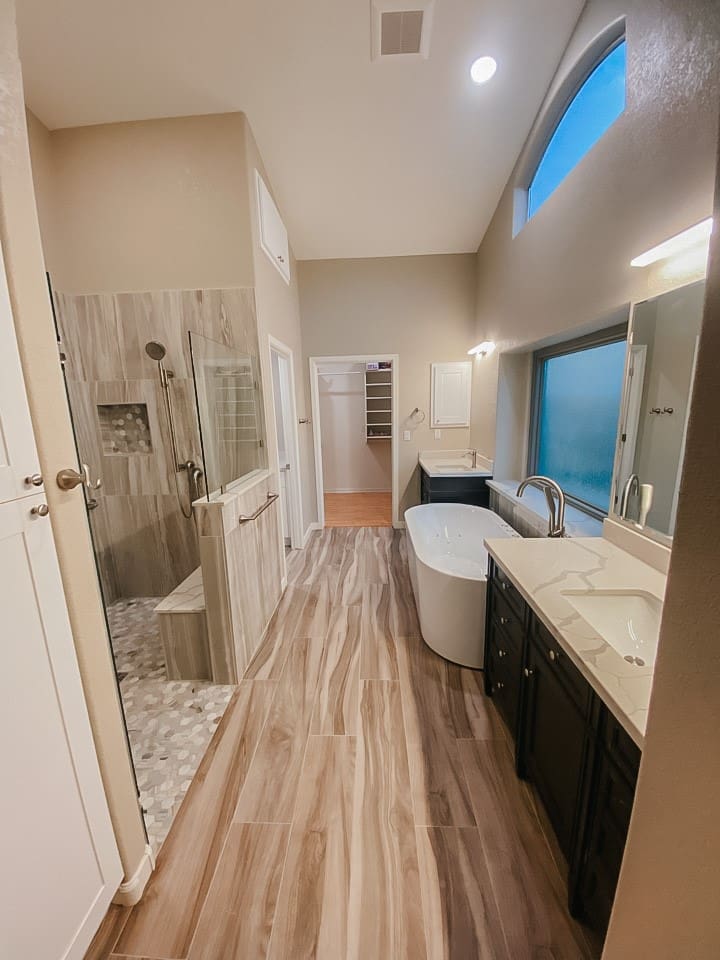 Modern bathroom with wood-look tile flooring, a glass-enclosed shower, a freestanding tub, dual vanities, and a view into an adjoining walk-in closet
