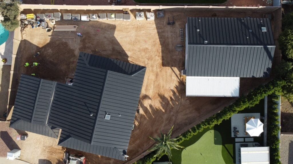 Aerial view of a construction site showing two buildings with dark metal roofs, surrounding dirt work areas, equipment, and workers along the perimeter