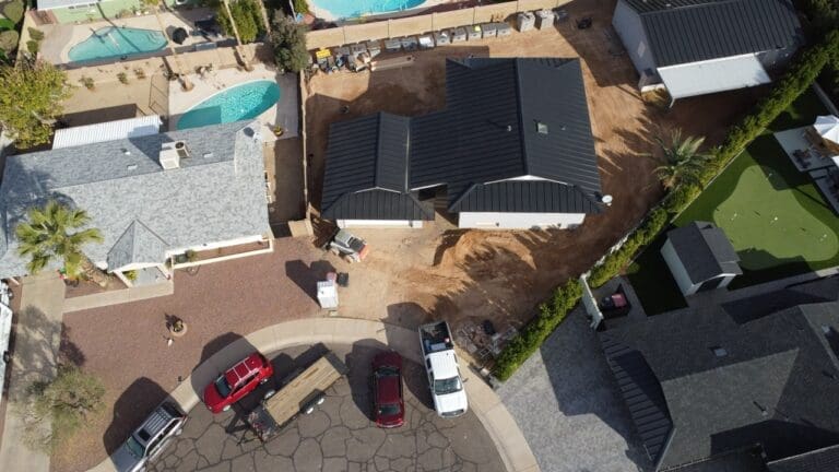 Aerial view of a residential construction site showing two buildings with dark metal roofs, surrounding dirt work areas, construction materials, and vehicles parked in the cul-de-sac
