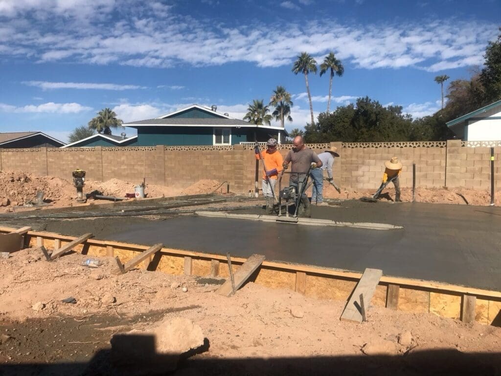 Construction crew pouring and smoothing fresh concrete for a foundation slab, surrounded by dirt mounds and a block wall fence