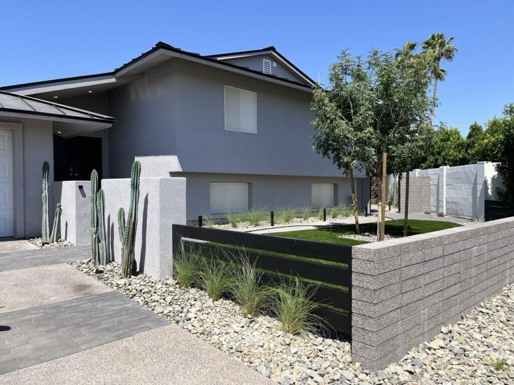 Front exterior of a modern home featuring clean stucco walls, desert landscaping with tall cacti and grasses, and a raised garden area enclosed by a contemporary block and metal fence.