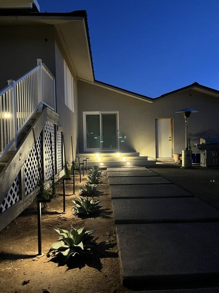 Outdoor walkway at night featuring pathway lights, agave plants, and illuminated steps leading up to a sliding door of a modern home