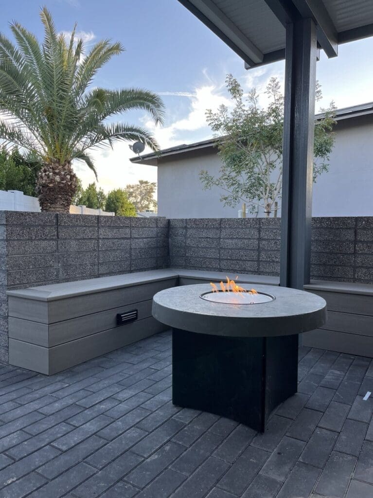 Outdoor patio area featuring a round concrete fire pit with visible flames, surrounded by built-in corner bench seating made of horizontal composite boards. A tall palm tree and a block privacy wall frame the space, with a covered patio roof overhead