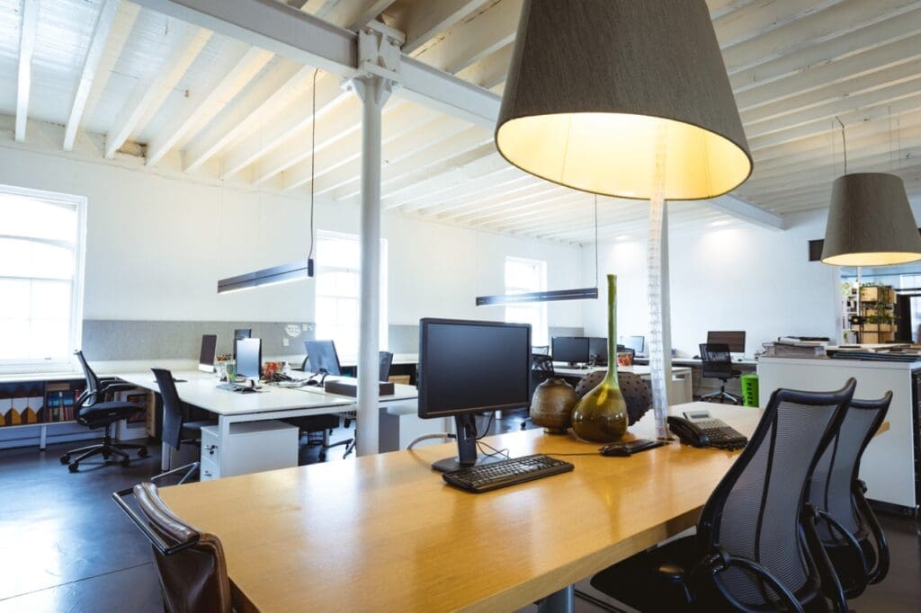 Bright modern office with wooden desks, computer monitors, office chairs, and large hanging lamps under a white-beamed ceiling