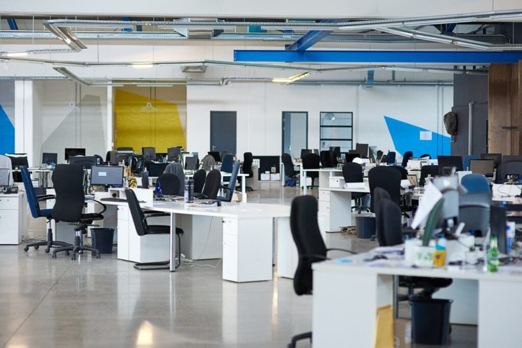 Spacious open-plan office with rows of desks, computers, and empty office chairs under exposed ceiling fixtures and bright lighting